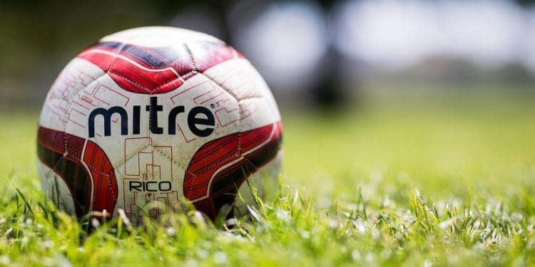 A close-up of a Mitre soccer ball resting on a grassy field with a blurred background.
