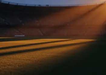 Empty floodlit stadium at dusk with packed stands, dramatic shadows, golden and red-white ambient lighting.
