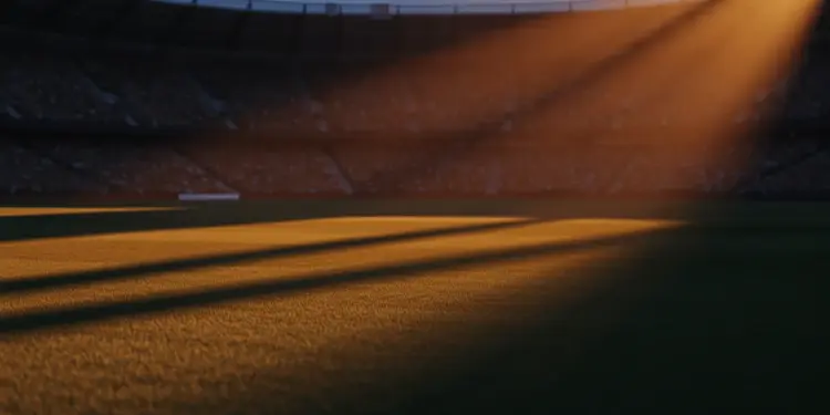 Empty floodlit stadium at dusk with packed stands, dramatic shadows, golden and red-white ambient lighting.