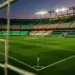 Floodlit stadium pitch in Algerian national colours—green and white—at dusk, with geometric pitch markings and goal net in sharp focus.