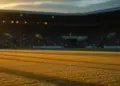 Floodlit football pitch at dusk with long shadows, packed stadium stands in background, golden-hour atmospheric lighting.