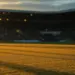 Floodlit football pitch at dusk with long shadows, packed stadium stands in background, golden-hour atmospheric lighting.