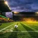 Floodlit football pitch at dusk with Australian-colour lit stands and a boot striking a ball in motion.