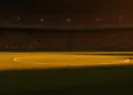 Floodlit stadium pitch at golden-hour kick-off with packed stands and dramatic shadow patterns cast across the grass.