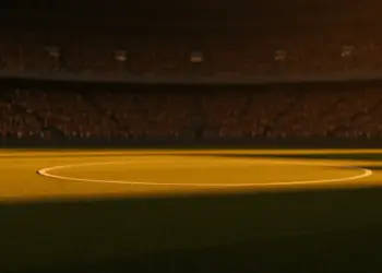 Floodlit stadium pitch at golden-hour kick-off with packed stands and dramatic shadow patterns cast across the grass.