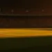 Floodlit stadium pitch at golden-hour kick-off with packed stands and dramatic shadow patterns cast across the grass.