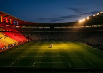 Floodlit stadium pitch at dusk with red-gold lighting and football on centre spot, Belgium national colour stands.