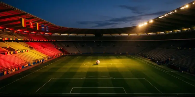 Floodlit stadium pitch at dusk with red-gold lighting and football on centre spot, Belgium national colour stands.