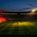 Floodlit stadium pitch at dusk with red-gold lighting and football on centre spot, Belgium national colour stands.