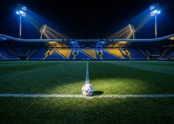 Floodlit stadium pitch with football on centre spot, blue and gold atmospheric lighting, sharp shadows, empty stands.