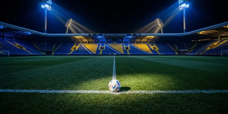 Floodlit stadium pitch with football on centre spot, blue and gold atmospheric lighting, sharp shadows, empty stands.