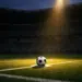 Floodlit football pitch at dusk with ball on penalty spot under dramatic single spotlight, empty stadium stands in shadow
