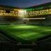Floodlit stadium pitch in green and gold tones with ball on centre spot, dramatic evening atmosphere.