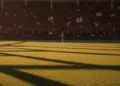 Floodlit stadium pitch with packed stands at golden hour, dramatic shadows across grass, no players visible.