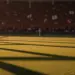 Floodlit stadium pitch with packed stands at golden hour, dramatic shadows across grass, no players visible.