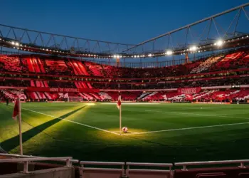 Floodlit stadium with red-draped stands, centre-spot ball, geometric shadows, dusk lighting.