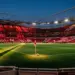 Floodlit stadium with red-draped stands, centre-spot ball, geometric shadows, dusk lighting.