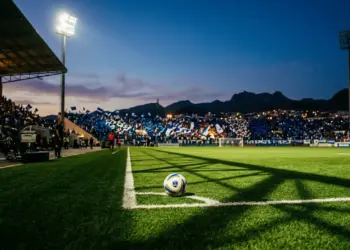Floodlit stadium pitch at golden hour with football on centre spot and Cape Verde-coloured crowd stands visible.