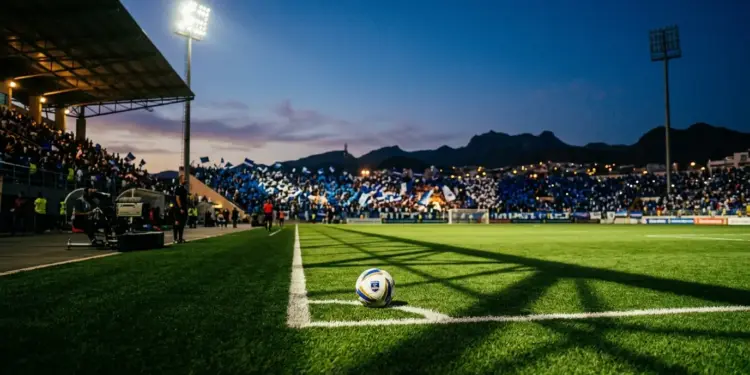 Floodlit stadium pitch at golden hour with football on centre spot and Cape Verde-coloured crowd stands visible.
