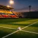 Floodlit stadium pitch at dusk with yellow-blue-coloured stand bunting, ball on centre circle, dramatic shadows and golden light.