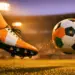 Football boot striking a ball mid-flight on a floodlit pitch at dusk, rendered in orange and green tones.