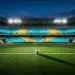 Floodlit stadium with cyan and gold stand sections, football on centre pitch under dramatic evening light.