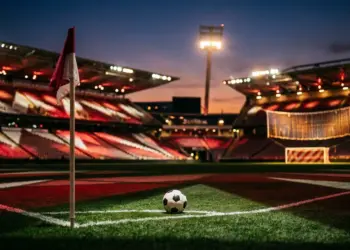 Floodlit stadium pitch at dusk with crimson and white lighting, corner flag visible, football on centre spot, empty stands in background.