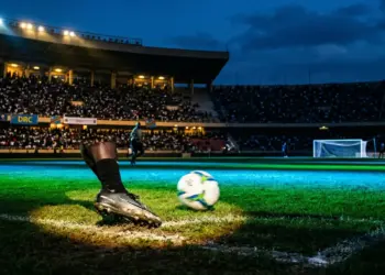 Floodlit football stadium with emerald and blue lighting, motion-blurred boot and ball, dramatic shadow patterns, goal net in silhouette.