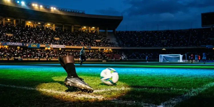 Floodlit football stadium with emerald and blue lighting, motion-blurred boot and ball, dramatic shadow patterns, goal net in silhouette.