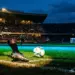 Floodlit football stadium with emerald and blue lighting, motion-blurred boot and ball, dramatic shadow patterns, goal net in silhouette.