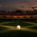 Floodlit football pitch at dusk with crimson and gold lighting, ball on centre spot, empty stadium stands.