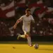 Football player in white kit striking ball at evening international match with flag-coloured stands in background.