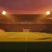 Floodlit stadium with packed stands at sunset, dramatic shadows on pitch, English and Ghanaian colour palette lighting.
