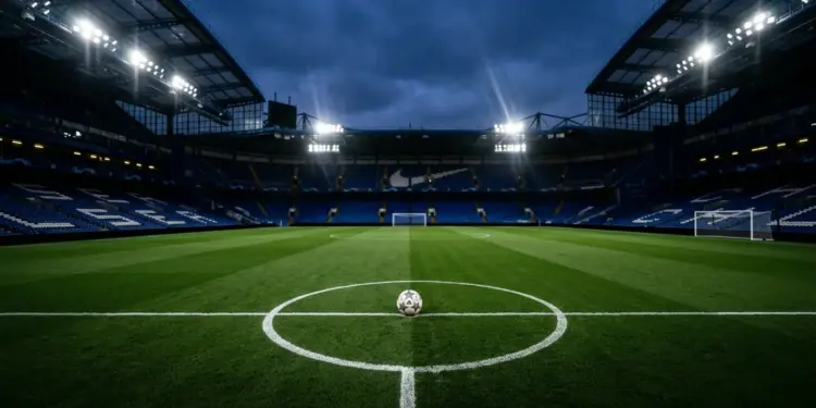 Floodlit stadium pitch with white markings, centre spot ball, royal-blue-tinted stands, dramatic shadow geometry, evening light.