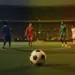 Floodlit pitch at dusk with players in unmarked kits, blue-gold and green-yellow-red coloured stadium lighting, football in foreground, shallow depth of field.