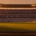 Floodlit stadium at dusk with blue and white spectators, dramatic shadows, golden-hour lighting, 16:9 landscape.