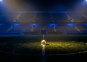 Empty floodlit football pitch with ball on centre spot, lit in blue and gold tones, dramatic stadium atmosphere.