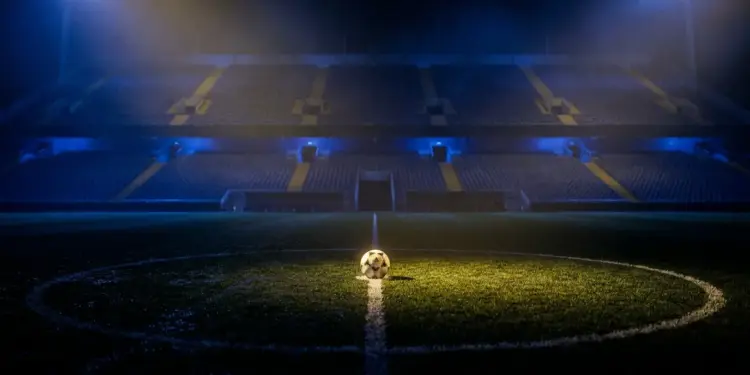 Empty floodlit football pitch with ball on centre spot, lit in blue and gold tones, dramatic stadium atmosphere.