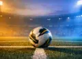 Match ball on centre spot of floodlit pitch at dusk, geometric shadows, stadium lighting, cool-blue and warm-gold atmospheric haze.