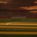 Empty floodlit stadium pitch at golden hour with packed stands, dramatic shadows, and cinematic lighting. No players visible.