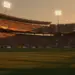 Floodlit stadium at golden hour with packed stands, dramatic shadows, and empty pitch ready for match.
