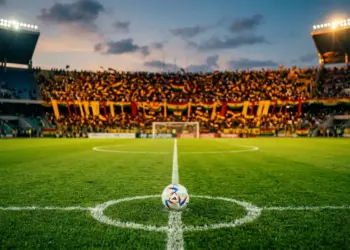 Floodlit stadium at dusk with centre pitch and gold-and-red draped stands under evening light.