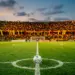 Floodlit stadium at dusk with centre pitch and gold-and-red draped stands under evening light.