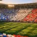 Stadium section with fans and flags in group colours, illuminated pitch with crowd anticipation.