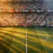 Overhead stadium view during evening kickoff: floodlit pitch, mixed-nation crowd colours, match ball on halfway line, dramatic shadows.