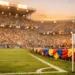 Floodlit stadium stands with flag-coloured bunting and packed crowd at sunset, pitch in foreground.