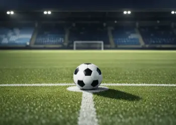 Floodlit football pitch with ball on penalty spot at dusk, blue and tartan stand sections in soft focus behind.