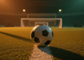 Floodlit football pitch at dusk with ball on centre spot, shadow patterns across turf, stadium lights golden-amber against cool shadows.