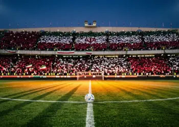 Floodlit football stadium at dusk with crimson and white stand sections, ball on centre circle under spotlight, shadows across pitch.