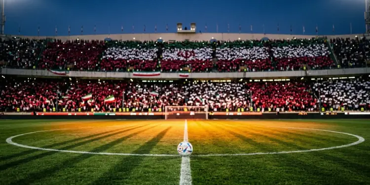 Floodlit football stadium at dusk with crimson and white stand sections, ball on centre circle under spotlight, shadows across pitch.